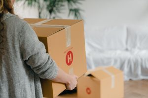 Back view of crop unrecognizable female in casual wear standing with square cardboard box in front of couch covered with crea