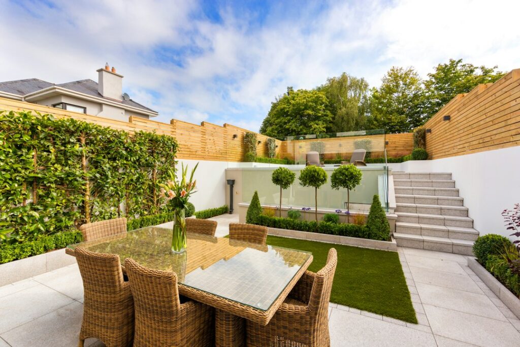 A modern small garden with a dining table and wooden fence, featuring greenery and glass boundaries.