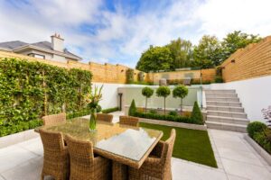 A modern small garden with a dining table and wooden fence, featuring greenery and glass boundaries.